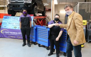 Steven Mitchell, Vice President of Mitchell Auto Group, stands next to two student recipients of the tool kits. Three blue tools kits are in the background, and a black pick up truck is on a car lift up in the air.i