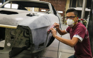 Automotive Collision Repair and Refinishing student poses for picture while wearing a protective face mask as he works on a car in the shop